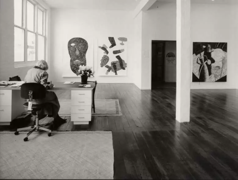 Sue Crockford at her desk in the Albert St. gallery, September 1985. On exhibition: works by Denys Watkins including The Sundays Were for Leisure (left) and Partially Dislocated (centre). (Photo courtesy of Art + Object and Sue Crockford).